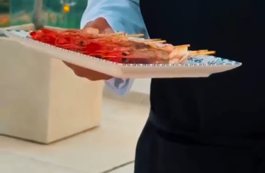 El chef llevando en la mano una bandeja de cerámica con brochetas de gamba, en la terraza de una casa particular con la costa al fondo