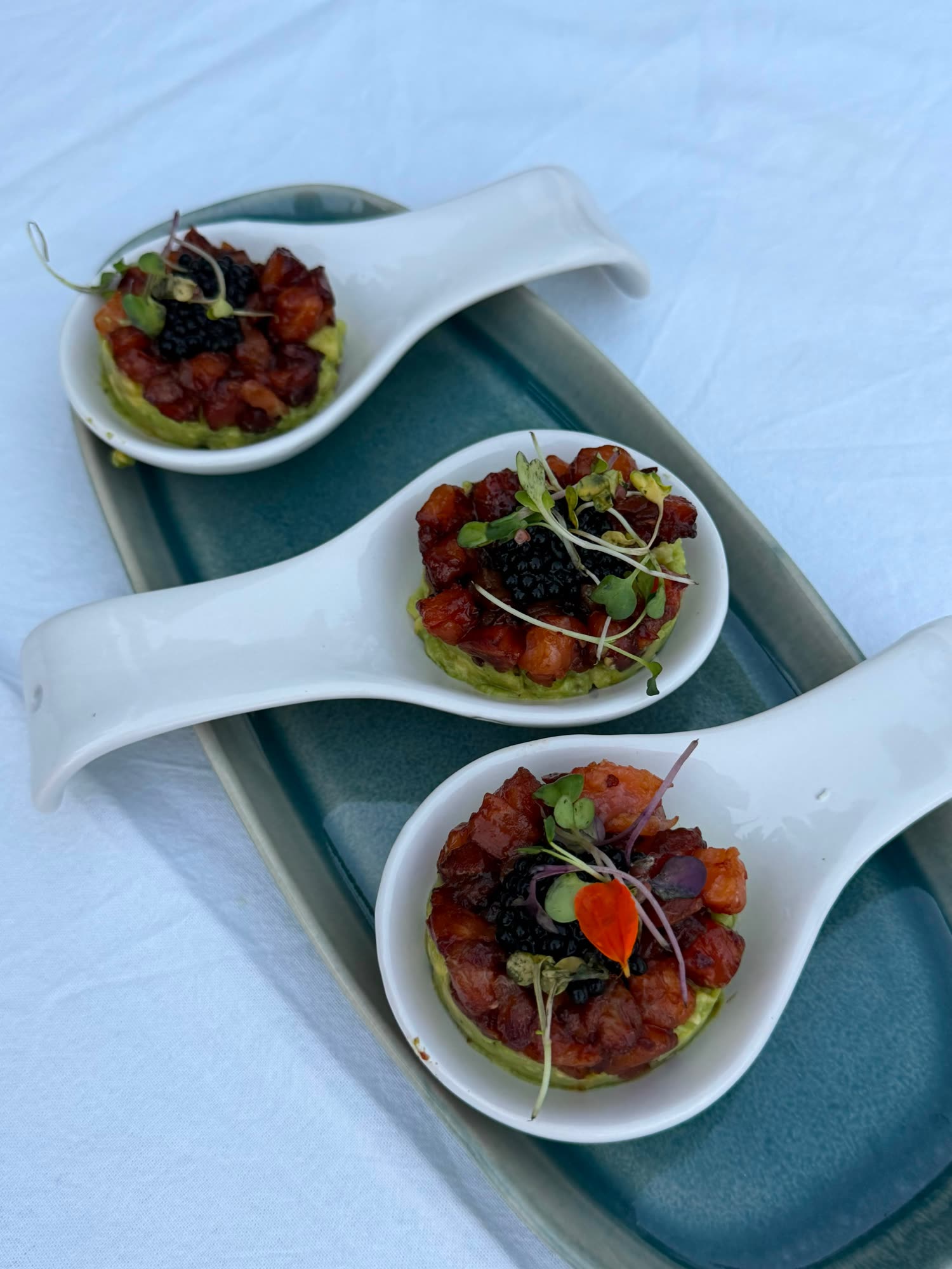 Tres cucharas de porcelana con tartar, perlas negras y microbrotes, sobre una bandeja de cerámica turquesa