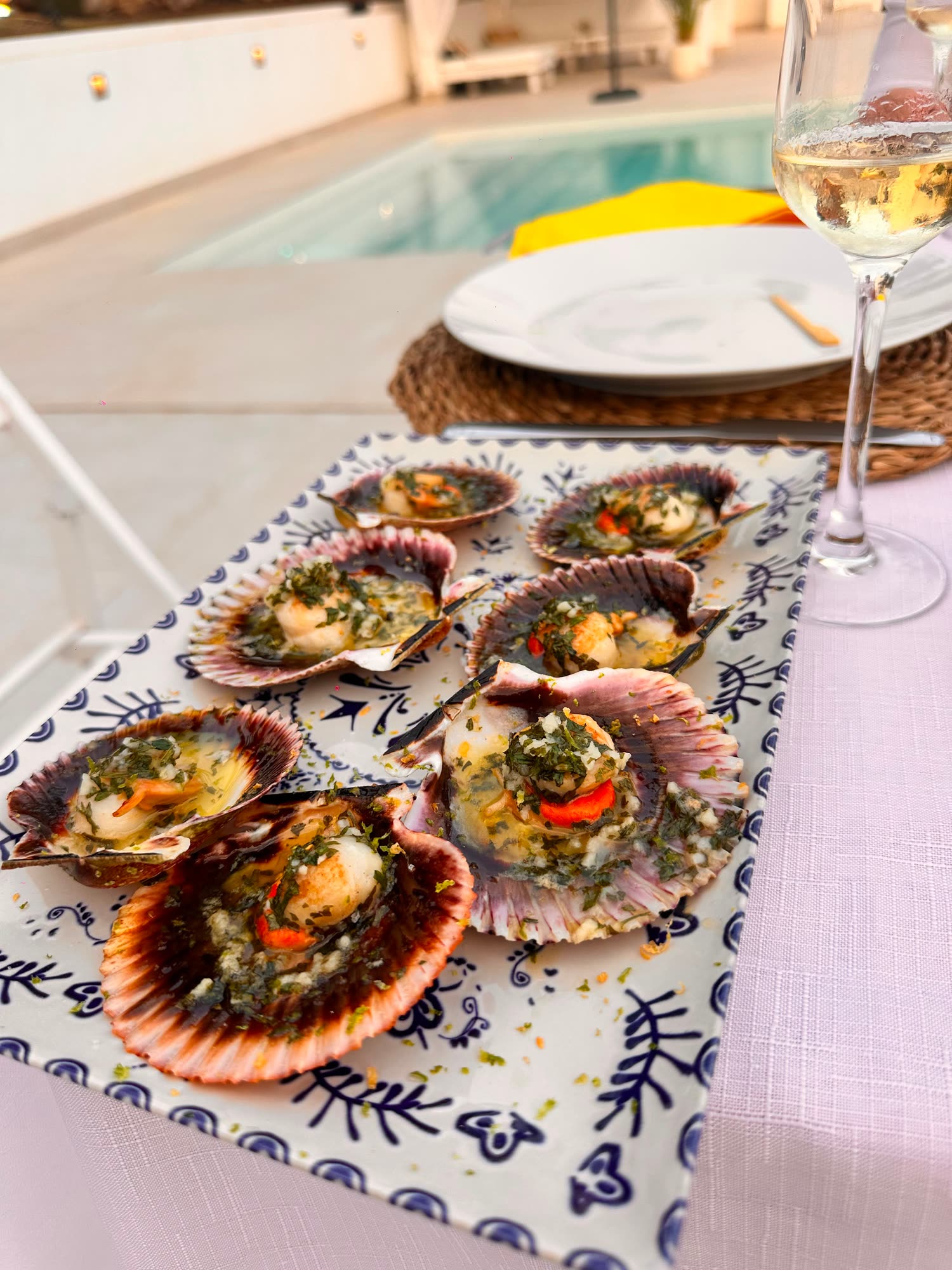 Zamburiñas gratinadas en su concha sobre una bandeja de cerámica azul, junto a una copa de vino blanco y la piscina de una casa particular al atardecer